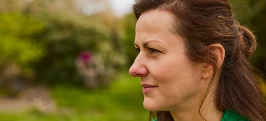 Nahaufnahme einer Frau im Profil, die im Freien in einem Garten sitzt. Sie trägt eine grüne Jacke und hat braune Haare. Ihr Gesichtsausdruck wirkt nachdenklich, möglicherweise aufgrund von Beschwerden durch eine trockene oder wunde Nase. Im Hintergrund sind unscharfe Bäume und Wiesen zu erkennen.