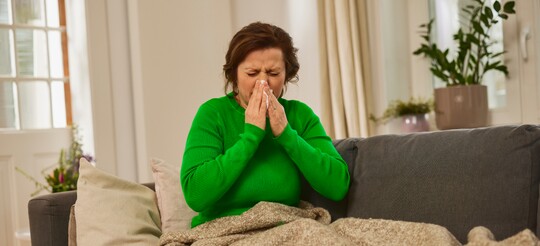 Frau mit Erkältung auf dem Sofa, eingehüllt in eine Decke und leidend unter einer verstopften Nase. Eine Frau in einem grünen Pullover sitzt auf einem Sofa, zugedeckt mit einer Decke, und schnäuzt sich in ein Taschentuch. Der Raum im Hintergrund ist hell und gemütlich eingerichtet.
