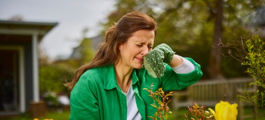 Eine Frau in einem grünen Hemd arbeitet im Garten und reibt sich die Nase mit einem Handschuh, möglicherweise aufgrund von Pollenallergie. Im Vordergrund sind Blumen zu sehen, und der Hintergrund zeigt Bäume und eine Bank im Garten.