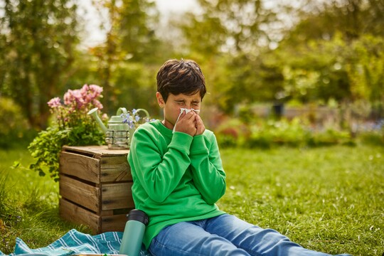 Ein Junge im grünen Pullover sitzt draußen auf einer Decke in einem Garten und schnäuzt sich die Nase. Neben ihm steht eine Wasserflasche, und im Hintergrund sind Pflanzen und Bäume zu sehen.