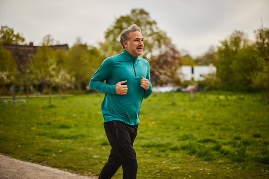 Ein Mann mittleren Alters joggt in einem grünen Langarmshirt durch eine grüne Landschaft. Im Hintergrund sind Bäume und Wiesen zu sehen. Er scheint sich wohlzufühlen, während er draußen trainiert.