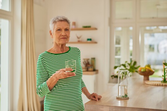 Ältere Frau mit grauen Haaren in gestreiftem Oberteil steht in einer modernen Küche und hält ein Glas Wasser in der Hand.