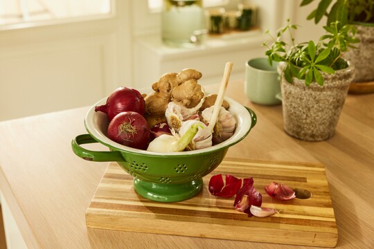 Frische Zutaten wie Zwiebeln, Ingwer und Knoblauch für natürliche Gesundheitsförderung. Eine grüne Schüssel mit roten Zwiebeln, Ingwer und Knoblauch steht auf einem Holzschneidebrett in einer modern eingerichteten Küche.