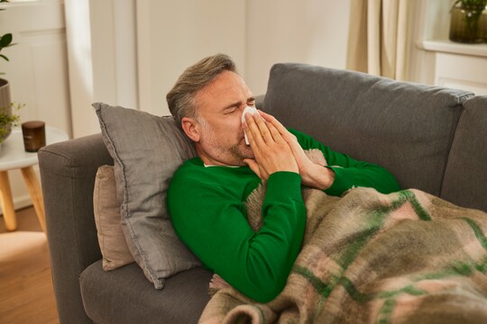 Ein Mann mittleren Alters in einem grünen Pullover liegt auf einem grauen Sofa. Er hält sich ein Taschentuch vor die Nase und niest. Er ist in eine Decke gehüllt und wirkt krank.