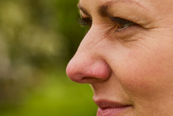 Nahaufnahme einer Frau im Profil, die im Freien in einem Garten sitzt. Sie trägt eine grüne Jacke und hat braune Haare. Ihr Gesichtsausdruck wirkt nachdenklich, möglicherweise aufgrund von Beschwerden durch eine trockene oder wunde Nase. Im Hintergrund sind unscharfe Bäume und Wiesen zu erkennen.