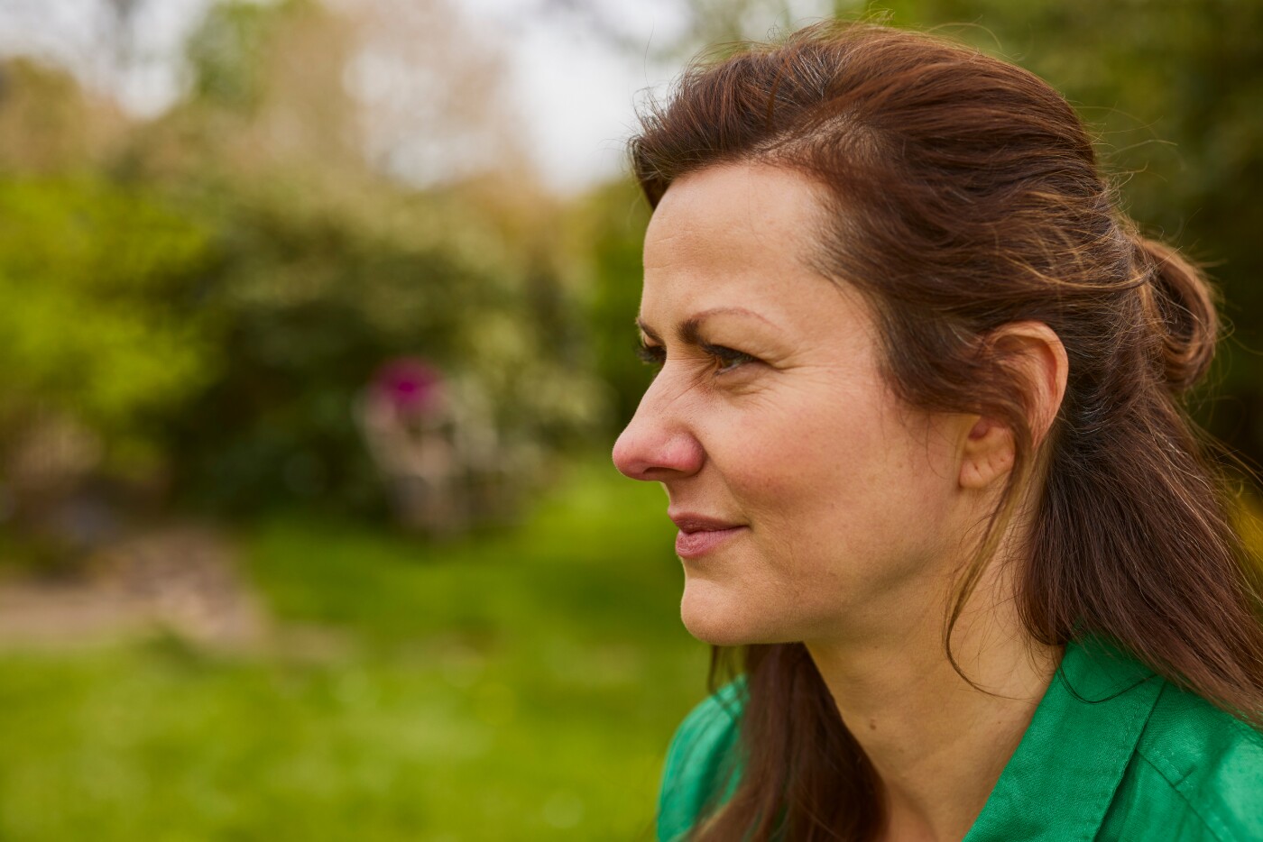 Nahaufnahme einer Frau im Profil, die im Freien in einem Garten sitzt. Sie trägt eine grüne Jacke und hat braune Haare. Ihr Gesichtsausdruck wirkt nachdenklich, möglicherweise aufgrund von Beschwerden durch eine trockene oder wunde Nase. Im Hintergrund sind unscharfe Bäume und Wiesen zu erkennen.